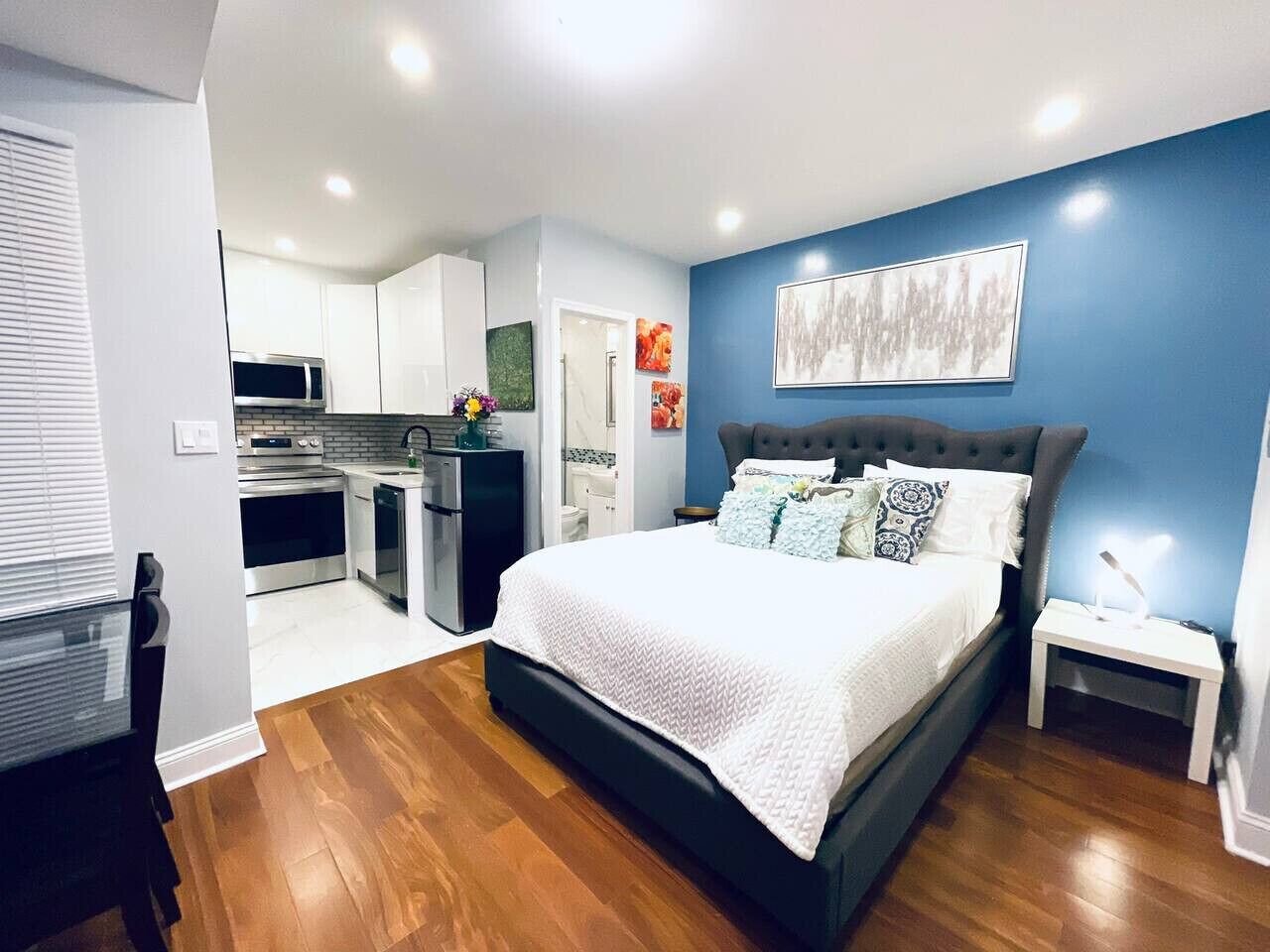 A modern bedroom with a large bed, a nightstand, and a view into the kitchen.