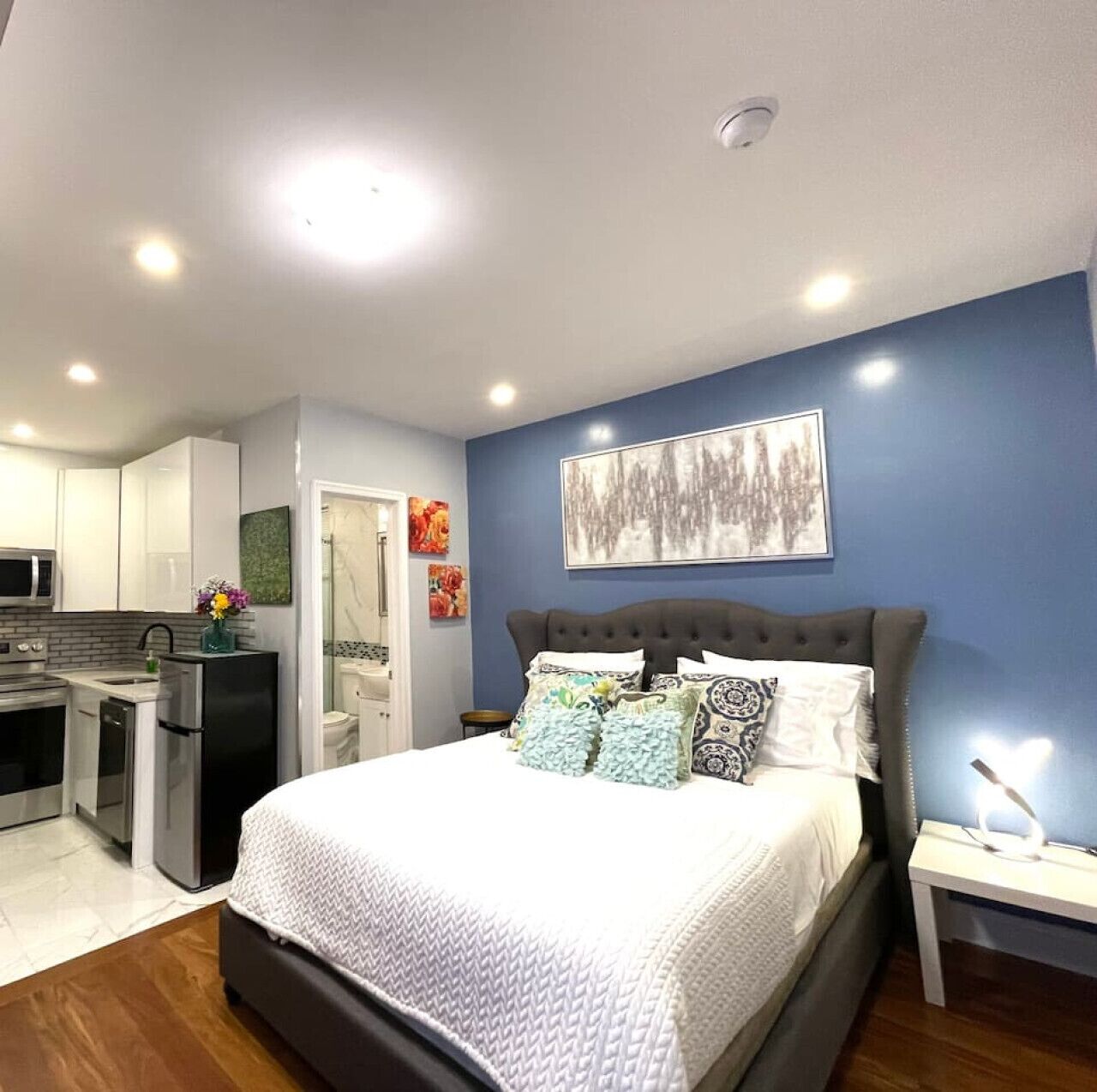 A bedroom with a bed, nightstand, and a view of the kitchen.