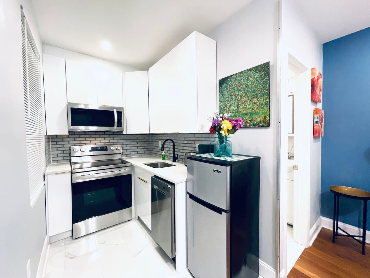 A modern kitchen with white cabinets, stainless steel appliances, a tiled backsplash, and a small refrigerator with a vase of flowers.