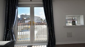 A room with a sliding glass door to a balcony, a small window with a clock, and a visible building with signage in the background.