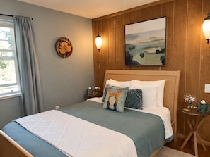 A bedroom with a wooden headboard, a bed with a quilt, a nightstand with a lamp, a wall clock, and a painting.