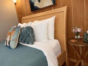 A bed with a wooden headboard, white bedding, and decorative pillows. There is a small round table with a vase of flowers and a candle.