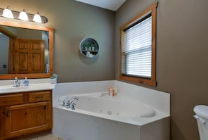 A bathroom with a large bathtub, a wooden vanity with drawers, a round shelf with toiletries, and a window with blinds. - Coyote Cabin (Branson)