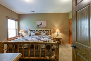 A bedroom with a wooden bed frame, a quilted bedspread, two bedside lamps, a window with blinds, and a door with a wooden frame. - Coyote Cabin (Branson)