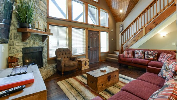 A spacious living room with a stone fireplace, a wooden staircase, and a large window with blinds. - Coyote Cabin (Branson)