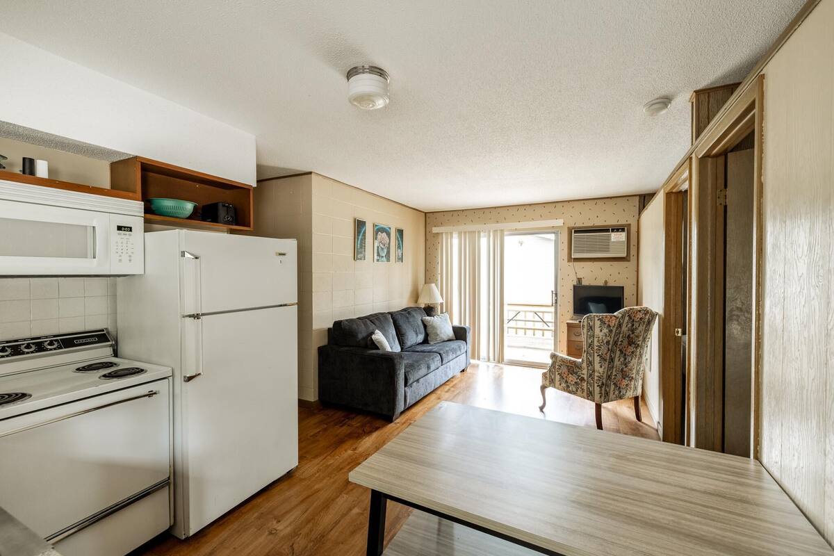 A compact kitchen with a microwave, stove, and refrigerator, a dark sofa, a wooden table, and a floral chair.