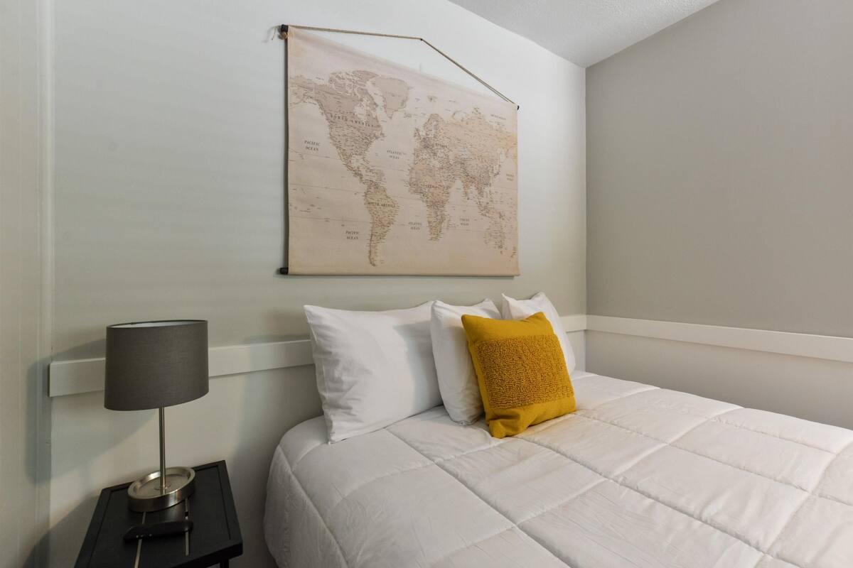 A neatly made bed with white bedding and a single yellow pillow, a black nightstand with a lamp, and a world map on the wall.