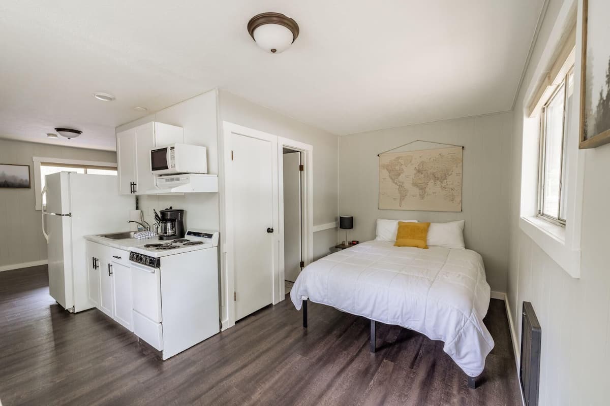 A compact room with a kitchenette, a bed with a world map on the wall, and a window.