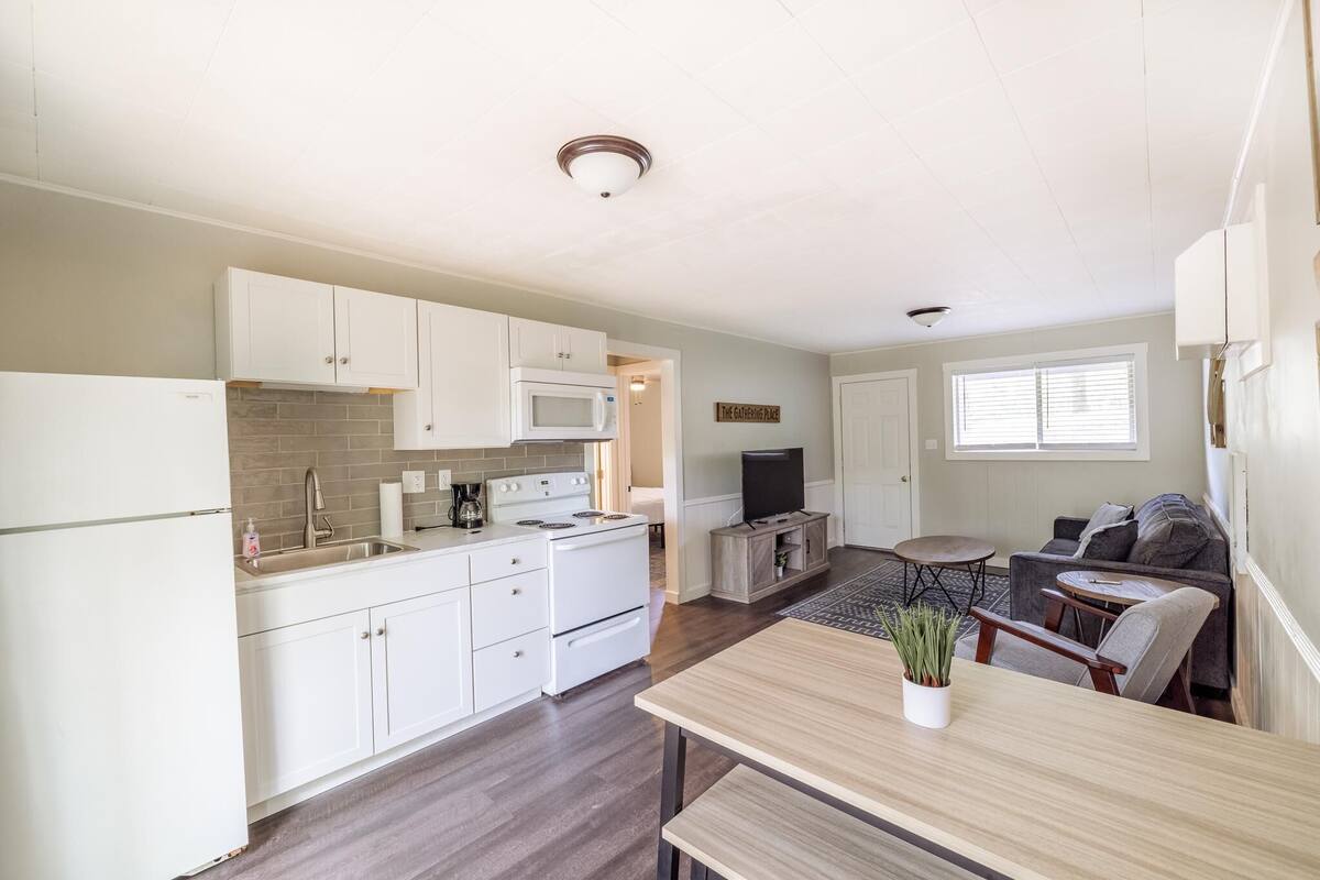 A compact kitchen with white cabinets, a microwave, and a stove. There is a dining area with a wooden table and chairs, and a small seating area with a couch and a TV.