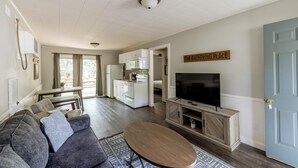 Un salon moderne avec un canapé gris, une table basse en bois et un téléviseur posé sur un meuble en bois. On y trouve également un coin cuisine avec des placards blancs et un réfrigérateur.