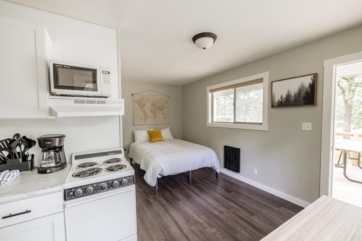 A compact kitchen with a microwave, stove, and coffee maker. A bed with a white comforter and a yellow pillow is against the wall. A map is hung on the wall, and a window provides natural light.