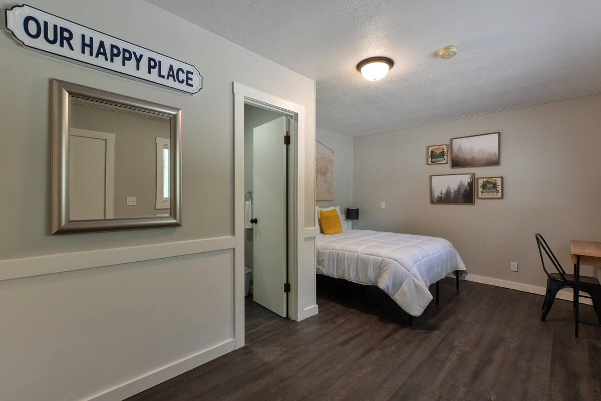A bedroom with a bed, a chair, a desk, a mirror, and a door. There is a sign on the wall that reads "OUR HAPPY PLACE".