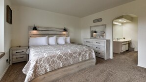 A bedroom with a large bed, a nightstand, a dresser, a vanity with a mirror, and a small archway leading to another room. - Farm Charm Condo (Branson)