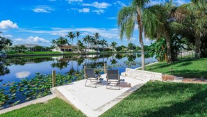 A serene lakeside setting with a concrete patio, two lounge chairs, and a small building in the background. - Incredible Lanai w/ Heated, Salt Pool, Extra-Wide Canal View, Southern Exposure! (Cape Coral)