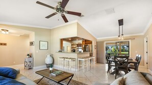 A spacious living room with a ceiling fan, a dining area with a view, and a kitchen with bar stools. - Incredible Lanai w/ Heated, Salt Pool, Extra-Wide Canal View, Southern Exposure! (Cape Coral)