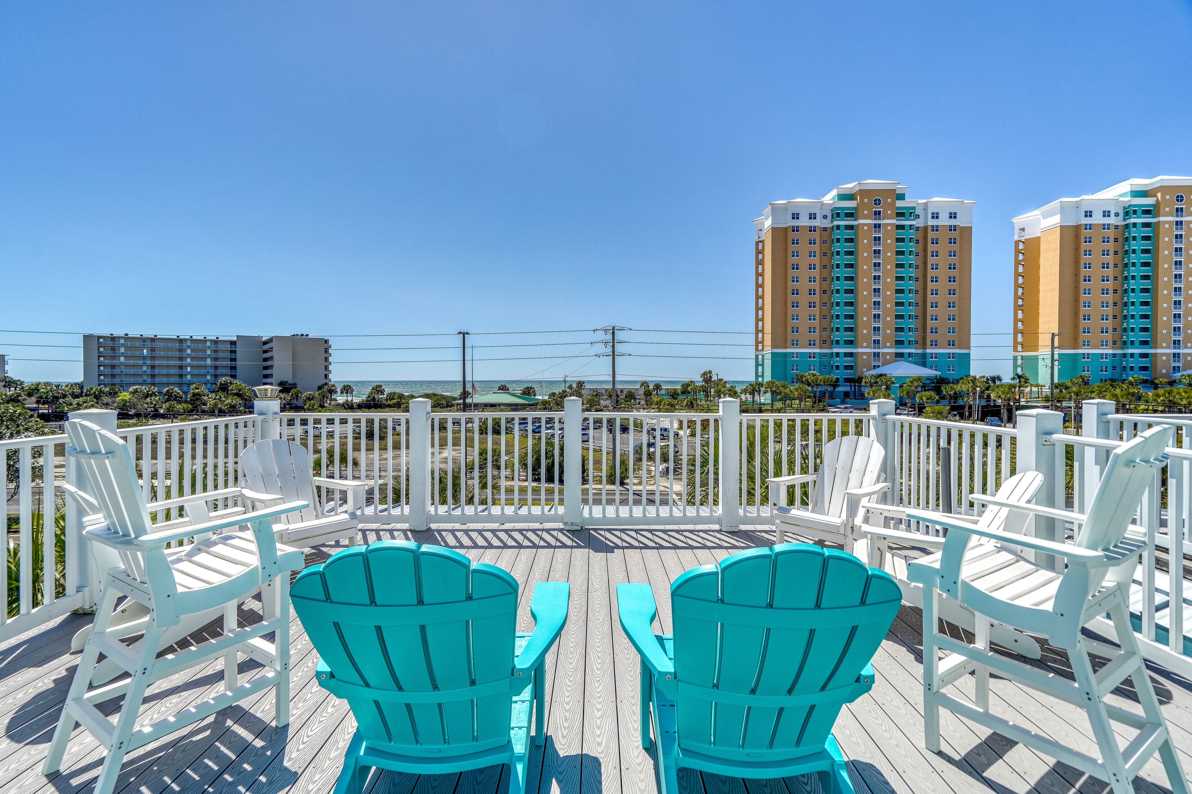 A deck with white and turquoise Adirondack chairs, a white fence, and a view of tall buildings in the background.
