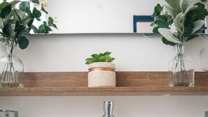 Une salle de bain avec un lavabo blanc, un porte-serviettes avec deux serviettes et deux étagères ornées de plantes et d’un petit récipient.