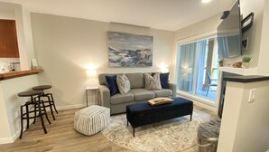 A modern living room with a grey sofa, striped ottoman, and a round rug. There is a kitchen area with a wooden countertop and stools. A painting is on the wall, and a TV is mounted above the counter. - Whistler Village Townhome (Whistler)