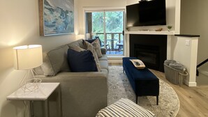 A modern living room with a grey sofa, a striped ottoman, a small table with a lamp, and a flat-screen TV mounted above a fireplace. - Whistler Village Townhome (Whistler)