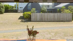 A view from a window with a wooden shelf holding five bottles, a small vase with dried plants, and a fenced backyard with a house and trees. - The Pirate's Rest, a tiny home by the beach. (Pauanui)
