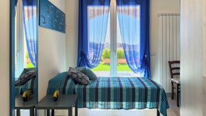 Un dormitorio con cama, una mesita y una silla. Se ve una ventana con cortinas azules y vistas al exterior con vegetación.