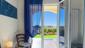 Una habitación con cama, una silla, una mesa con lámpara, y vista a un jardín verde y edificios a través de una puerta de vidrio.