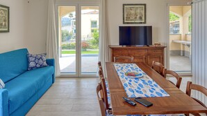 Una habitación con un sofá azul, una mesa de comedor de madera con mantel estampado de peces, un televisor sobre un mueble de madera y vistas a un jardín a través de puertas de cristal.