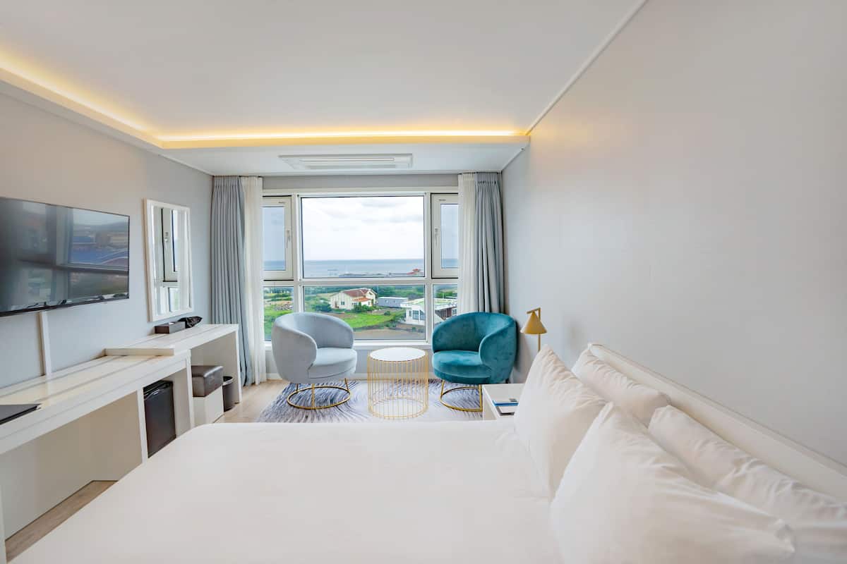 A hotel room with a bed, a desk, a TV, two chairs, a small table, and a view of the sea.