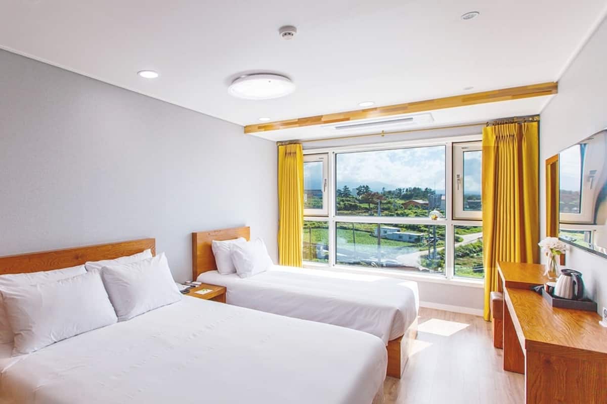 A hotel room with two beds, a wooden headboard, a desk with a chair, and a large window with curtains.