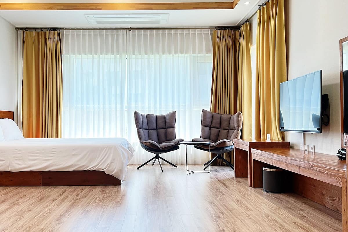 A hotel room with a bed, two armchairs, a TV, and a large window with curtains.