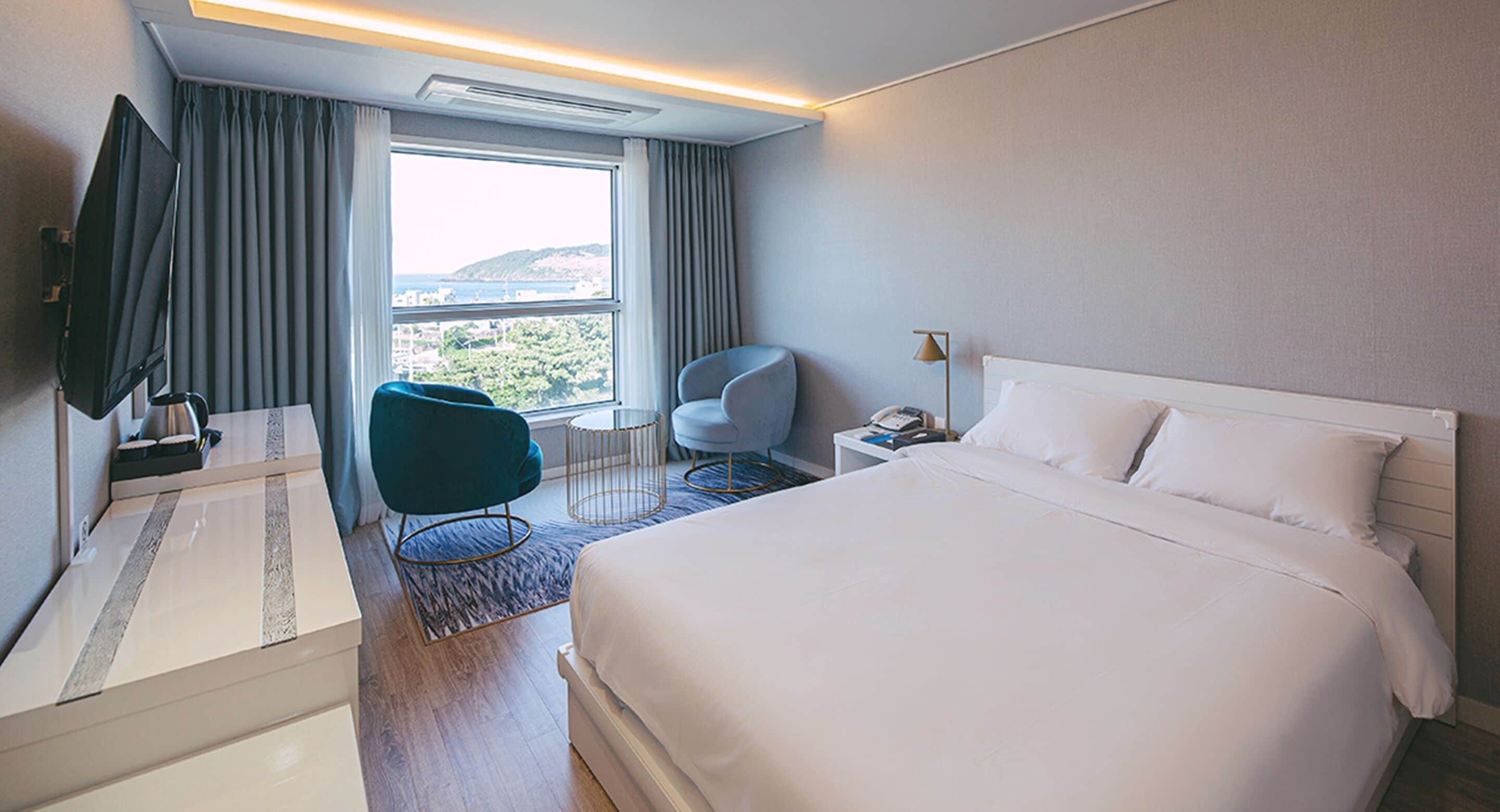 Ein Hotelzimmer mit einem großen Bett, einem Schreibtisch mit Flachbildfernseher, einem Sessel, einem Nachttisch und Blick auf die Stadt.