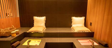 A spa room with two cushioned seating areas, a small sink, and a wooden panel on one side.