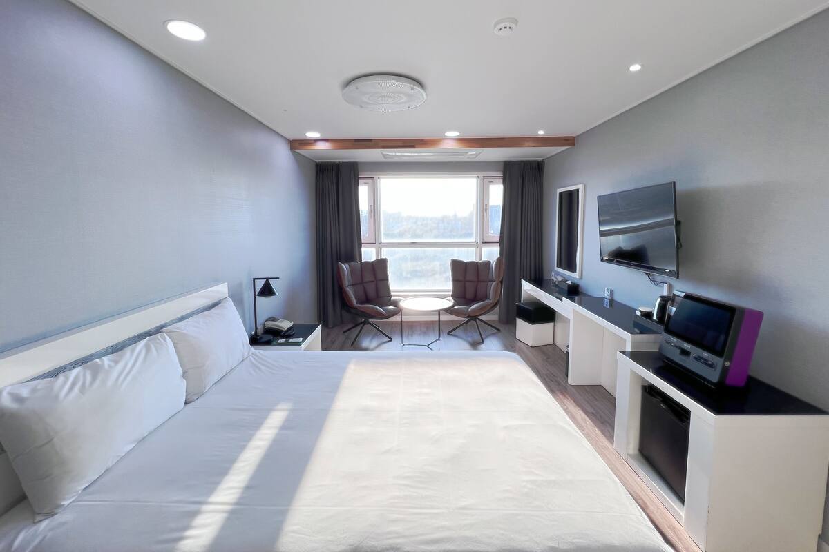 A hotel room with a large bed, a desk with a flat-screen TV, a chair, and a window with a view.
