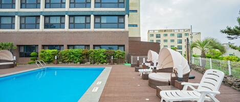 A swimming pool with lounge chairs and umbrellas, surrounded by a wooden deck and a building with multiple windows.
