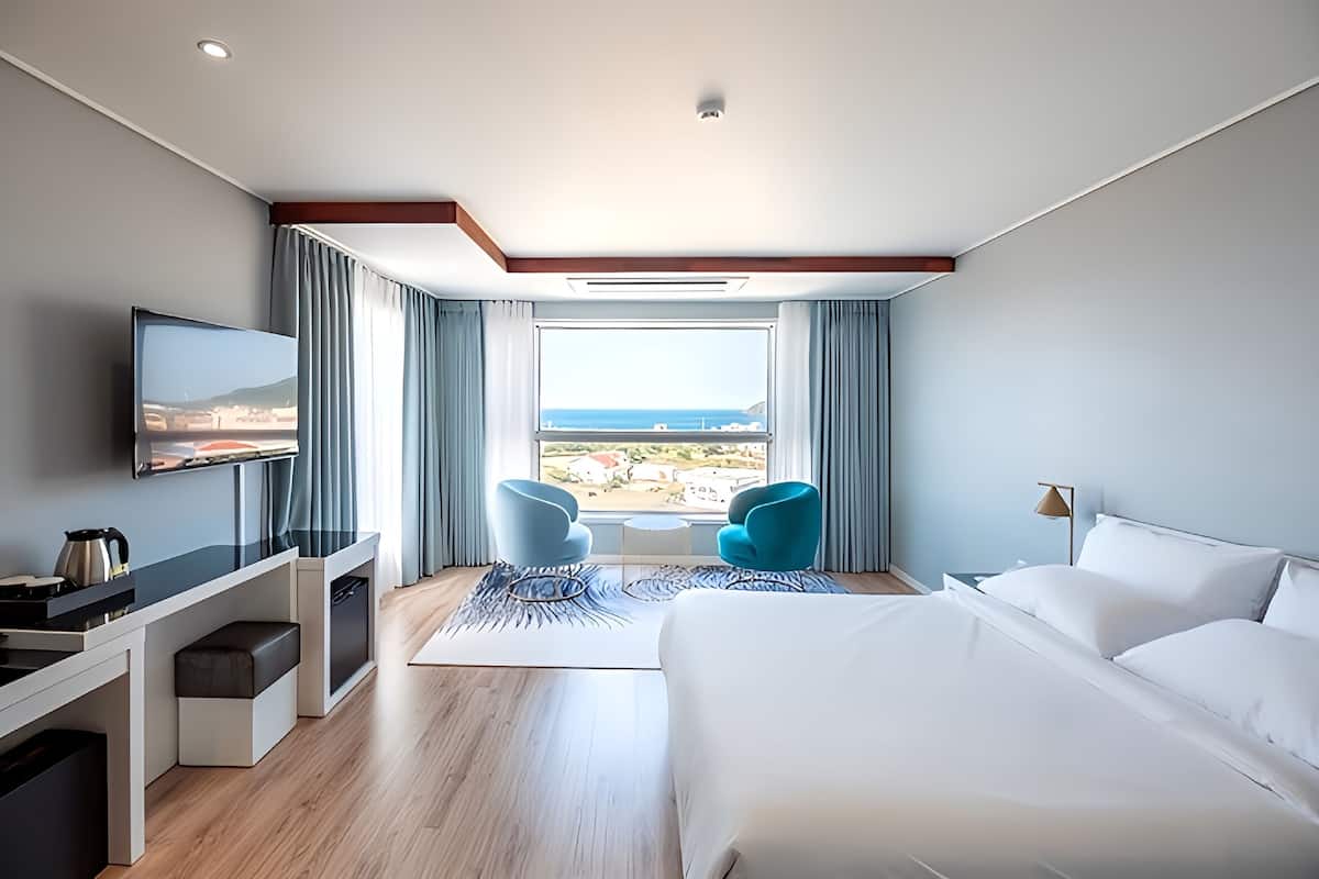 A modern hotel room with a large bed, a desk with a flat-screen TV, two blue chairs, a view of the sea, and a large window.