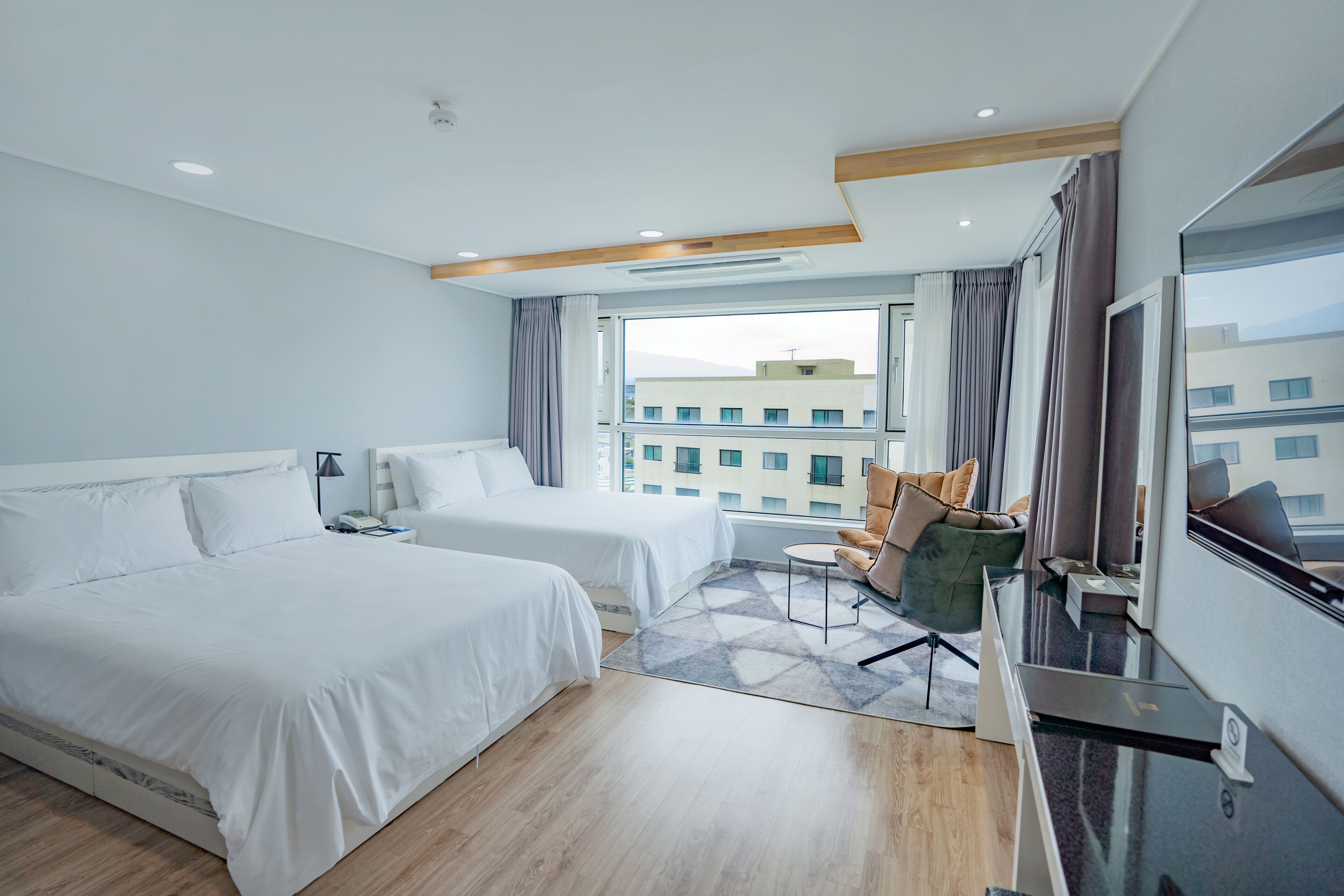 Ein Hotelzimmer mit zwei Betten, einem Schreibtisch, einem Stuhl und einem großen Fenster mit Blick auf die Stadt.