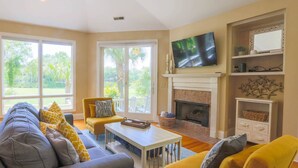 A spacious living room with a fireplace, a television mounted above it, and a large window providing a view of the outdoors. - Gorgeous Natural Light, Vaulted Ceilings & Fairway Views.  Back Deck & Screened Porch. (Kiawah Island)