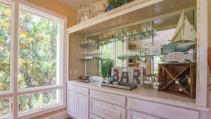 A kitchen with a large window, white cabinetry, and a bar area with glass shelves and various items. - Gorgeous Natural Light, Vaulted Ceilings & Fairway Views.  Back Deck & Screened Porch. (Kiawah Island)
