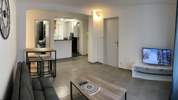 A modern living room with a sofa, coffee table, and TV. There is a kitchen area with a dining table and chairs. The room has a large clock on the wall and a sliding door leading to another room. - Joli T2 Climatis? Nimes Proche Gare et Centre Ville (Nîmes)