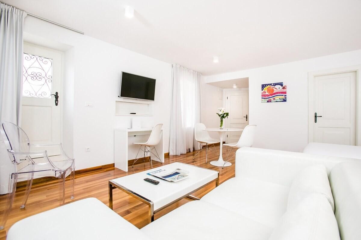Un salon moderne et bien éclairé, avec un canapé blanc, une table basse et des chaises transparentes. Un téléviseur à écran plat est fixé au mur, et une petite table avec un vase de fleurs se trouve à proximité.