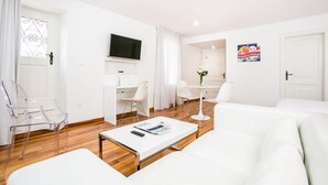 A modern, well-lit living room with a white sofa, a coffee table, and clear chairs. A flat-screen TV is mounted on the wall, and there is a small table with a vase of flowers. - Deluxe Studio with Balcony 302 Kovačića Street 20 (Split)