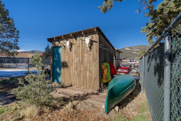 A wooden shed with a green door, a cow skull mounted on the wall, and a green canoe leaning against a fence.