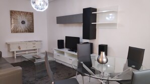 Un salon moderne avec une table à manger en verre, des chaises noires, un meuble blanc, un téléviseur à écran plat et un vase décoratif.