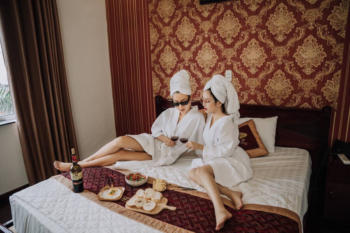 A room with a patterned wallpaper, a bed with a red and white bedspread, a bottle of wine, and a tray with bread and a bowl of fruit.