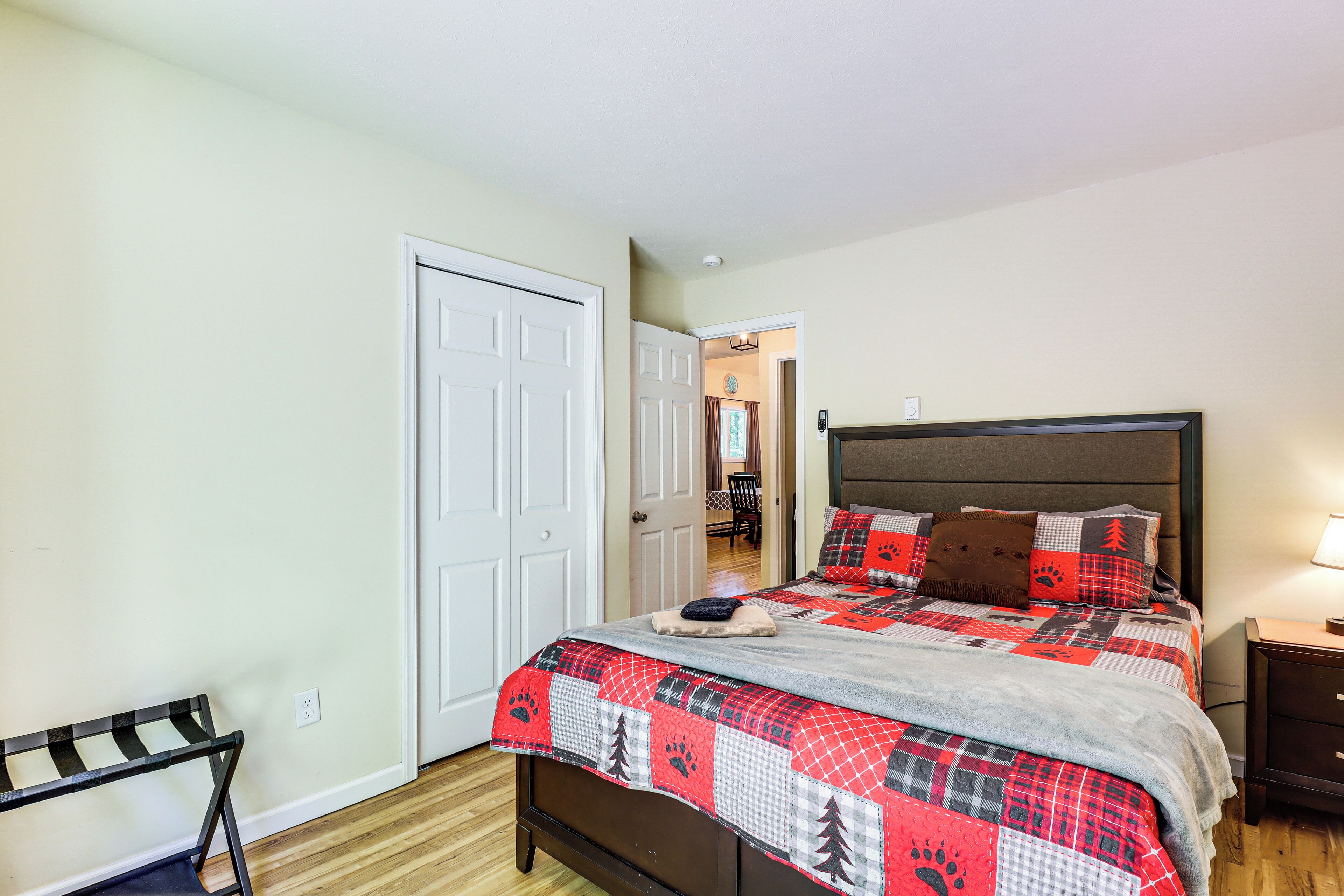Quarto com cama, um banquinho, uma porta e uma mesa de cabeceira.