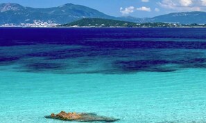 A coastal scene with clear turquoise waters, rocky formations, and distant mountains.