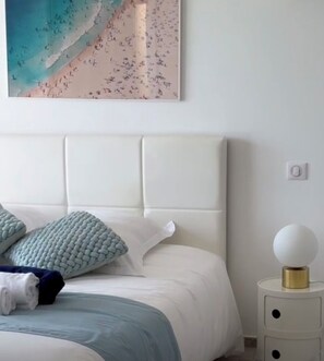 Un lit bien fait, avec une tête de lit blanche, une couverture bleue et un coussin décoratif. On y trouve une petite table de chevet sur laquelle se trouve une lampe blanche et un objet rond.