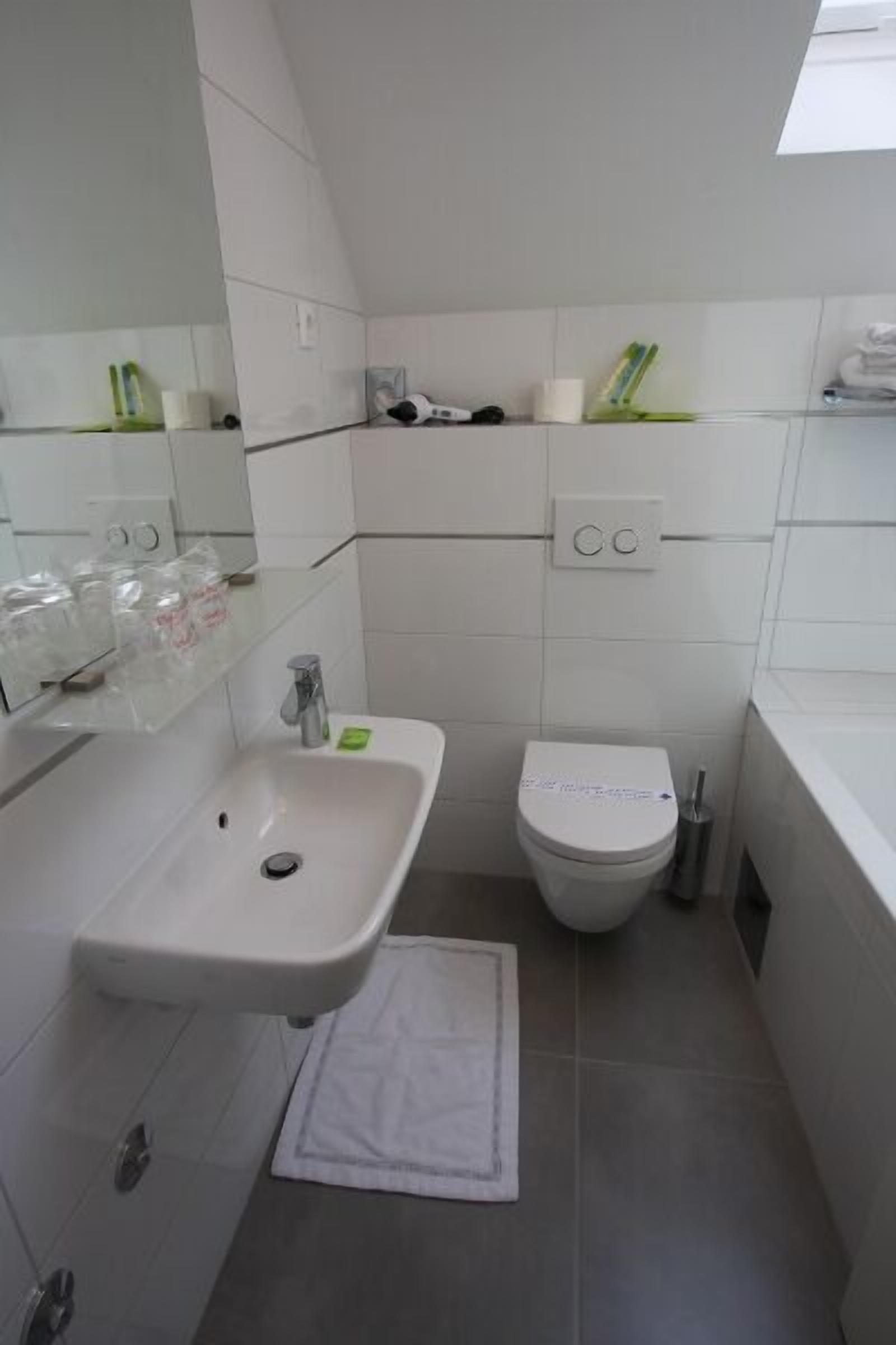A small, modern bathroom with a white sink, toilet, and a mirror above the sink. There are toiletries on the sink and a towel on the floor.
