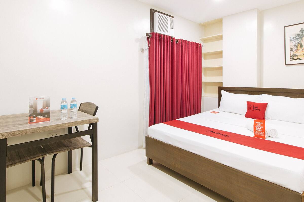 A hotel room with a wooden bed, a red curtain, a table with water bottles, and a framed picture on the wall.
