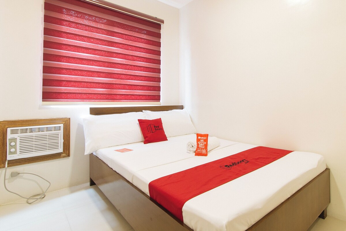 A hotel room with a bed, a window with red blinds, a white pillow, a red pillow, a red blanket, and a white bedside table with a radio.